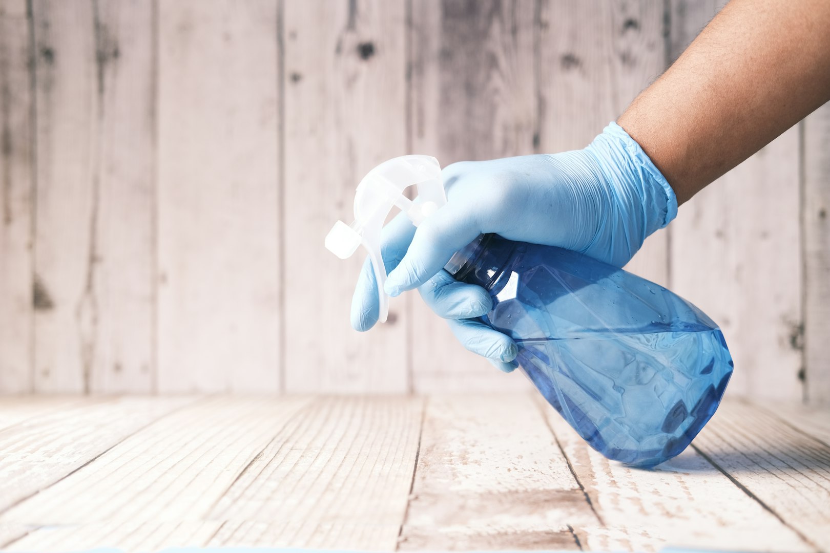 A man wearing yellow cleaning gloves, a white shirt, and a blue apron is cleaning a window with a spray bottle and a cloth. He is smiling and appears focused on the task. Indoor plants and a soft couch are visible in the background.